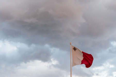 Malta flag. Malta flag on a flagpole waving. Bad weather clouds as a background during sunset hour. Horizontal.の写真素材