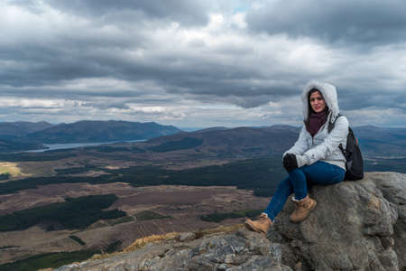 Woman with fluffy white jacket and hiking shoes posing on a mountain with mountains and lakes in the background, on a cloudy day at Nevis Range, Scottish Highlandsの写真素材