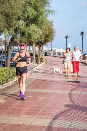 16th September 2016 Sliema Malta. Middle aged 40-50 woman jogging with sports clothes and violet running shoes along the front. two people at the background walking with dog.のeditorial素材