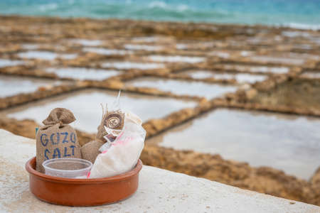 Salinas. Salt Pans or salters at the background with salt bags ready for sale in a basket or bowl outdoor. Hand harvest salt in Gozo Island Marsalforn. space for text.のeditorial素材