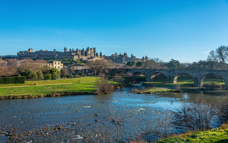 Carcasonne cityscape panoramic view over aude river in winter under blue sunny sky. Medieval fortified stone wall on hill top, Pont Vieux old stone bridge crossing the Aude river on the right.のeditorial素材
