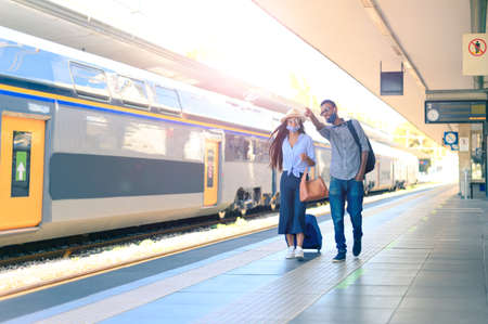 Couple on the platform waiting for the train - Beautiful woman and handsome man in medical masks walking and having trip during pandemic - New normal lifestyle conceptの写真素材