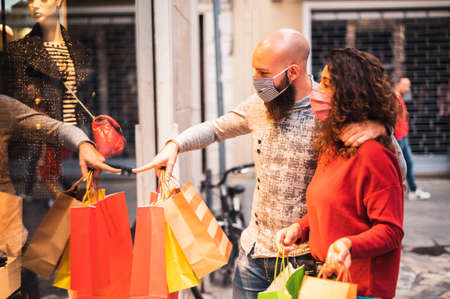 Pretty young man pointing to shop window to show clothing item his likes to his girlfriend - Beautiful young couple enjoying in shopping, having fun together, with the face mask - Consumerism, love, dating, new normal, lifestyle conceptの写真素材