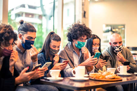 Multiracial friends with face mask holding smartphones sitting at table drinking cappuccino at cafe shop in the covid-19 time - People having fun together eating cakes and using mobile smart phone at cafe bar - New normal friendship conceptの写真素材
