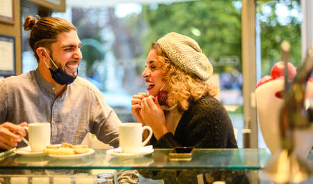 Beautiful happy loving couple with face mask enjoying breakfast in a cafe in the coronavirus time -Two smiling friends drink capucino and eat cookie at cafe shop - Love, dating, food, new normal lifestyle conceptの写真素材