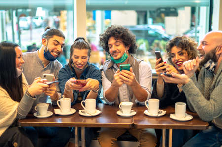Two man looking in phone over the shoulder of girl friend and trying to see what is on the screen, in the cafe shop - Group of happy friend with the face mask talking laughing and enjoying their time using smartphone and drink capuccino in the coronavirus time - New normal lifestyle conceptの写真素材