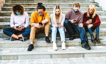 Multiracial friends with face masks using smart phones sitting on the stairs in streets city - Young millennial people watching content on social media networks - New normal lifestyle concept - Cold filterの写真素材