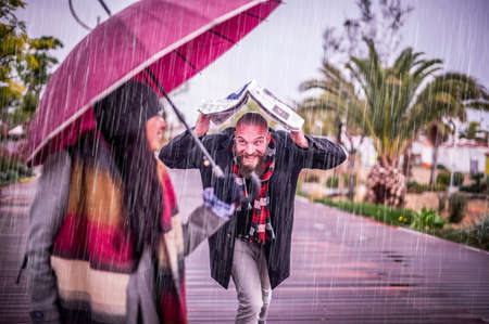 The loving couple in the rain - Beautiful girl holding umbrella while her boyfriend runs to her under the rain under a newspaper - She is turning and looking at him with true delight and sincere loveの写真素材