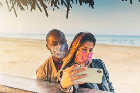 Multiracial millennial friends taking selfie smiling behind face masks at bar on the beach - Happy friendship and new normal concept with young people having fun togetherの写真素材
