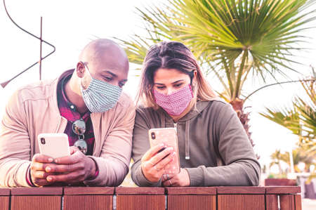Interracial couple with face mask watch a smartphone together on holidays - Multiracial trendy friends next to each other with telephone in the hands outdoors - New normal lifestyle conceptの写真素材