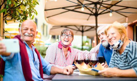 Senior couples taking selfie at restaurant bar with face masks - New normal lifestyle concept with happy people having fun together outdoors in covid-19 time - Focus on woman with glassesの写真素材
