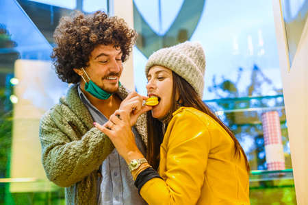 Beautiful happy couple feed each other in pastry shop - The young man feeds a macarons to his girlfriend - New normal lifestyle concept in covid-19 timeの写真素材