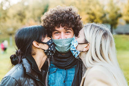 Love triangle - Two young attractive women with face masck kissing man outdoors in coronavirus time...