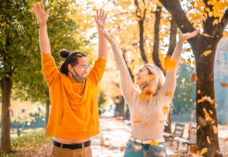 Happy smiling couple with face mask down, throwing autumn fall orange red leaves in park forest...