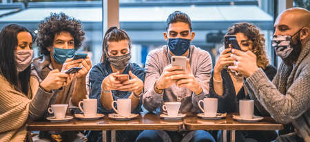 Group of young multi-ethnic people using smartphone sitting in a coffee shop with face mask protection - Multiracial people holding smart mobile phones and watching their screen in pandemic timeの写真素材