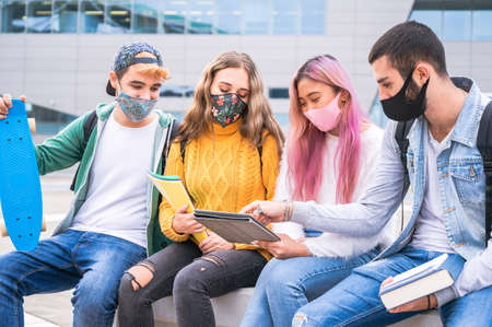 Multiracial teenage friends wear face mask sitting on tha bench outside the school - New normal lifestyle concept with young students studing together near to the campus outdoorsの写真素材