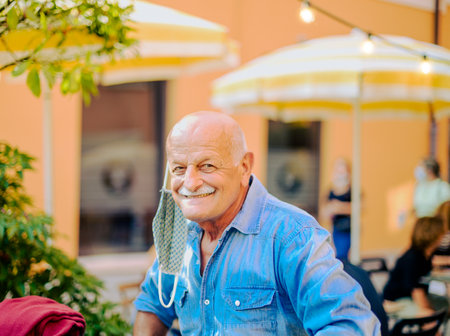 Elderly man with open protective face mask sitting in a bar - Joyful senior man see at the camera - Concept of health care and safety measures in pandemic timeの写真素材