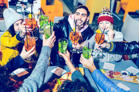 Friends toasting colored cocktails and drinking outside at night in the cocktail bar wearing a face...