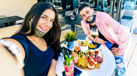 Happy smiling couple with face mask down taking selfie during happy hour at bar - Fashion man and young woman having fun at cocktail bar - Fashion concept with boyfriend and girlfriendの写真素材