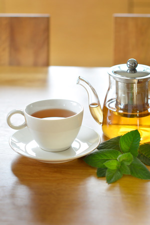 White ceramic cup and glass teapot of herbal tea with fresh mint twig on wooden tableの写真素材