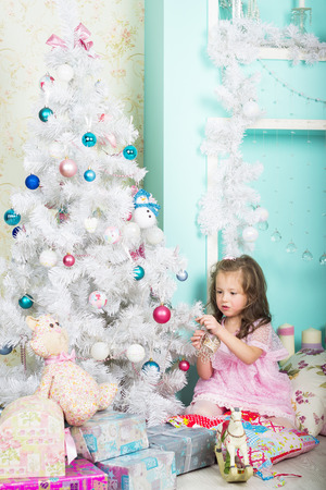 Waiting for Christmas: a beautiful little girl with a winter makeover in a pink dress decorates the new year tree on the background of stylish new year decorations in the style of high keyの写真素材