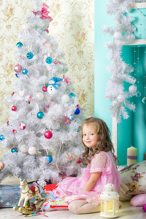 Waiting for Christmas: a beautiful little girl with a winter makeover in a pink dress decorates the new year tree on the background of stylish new year decorations in the style of high keyの写真素材