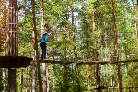 Teenage girl goes on hinged trail in extreme rope Park in summer forest. High-altitude climbing training of child on adventure track, equipped with safety straps and protective helmetの写真素材