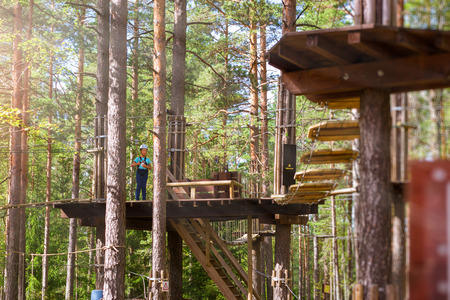 Teenage girl goes on hinged trail in extreme rope Park in summer forest. High-altitude climbing training of child on adventure track, equipped with safety straps and protective helmetの写真素材