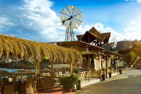 Decorative windmill with white propeller installed on thatched roof. Tables with thatched umbrellas at beach cafe on seafront. Livadi beach in sea bay of resort village Bali. Crete, Greeceのeditorial素材