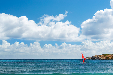 Windsurf with red sail rides on sea on background rocky shore. View from sunny Livadi beach in resort village Bali. Rethymno, Crete Greece. Extreme water sports as active recreation on Sunny vacationsの写真素材
