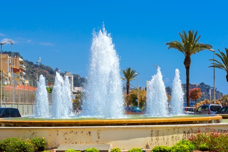 Blanes, Spain - 31 may, 2018: Round fountain among the road junction. Water jets hit up. Architecture of Spanish beach resort Blanes in summertime. Costa Brava, Catalonia. Flower bed in city squareのeditorial素材