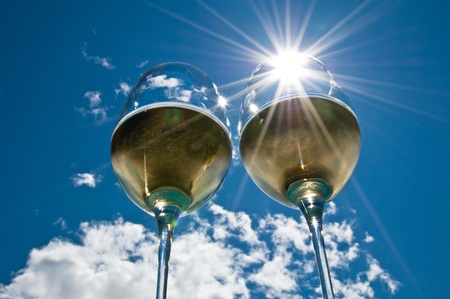 closeup of two wineglasses with a sun burst filled with white wine side by side with bright blue sky & clouds in the backgroundの写真素材