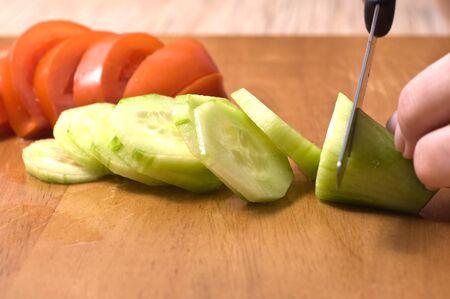 Hands slicing cucumber  on wooden board. Tomatoe in background.の写真素材