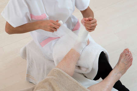 Nurse Taking Care of senior woman in her retirement home bandaging ankle and footの写真素材
