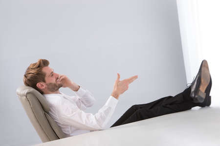 Young businessman in office sitting with feet on desk talking on cellphone.の写真素材