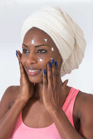 Portrait of beautiful young AfroAmerican woman wrapped in towel spreading cream on her faceの写真素材