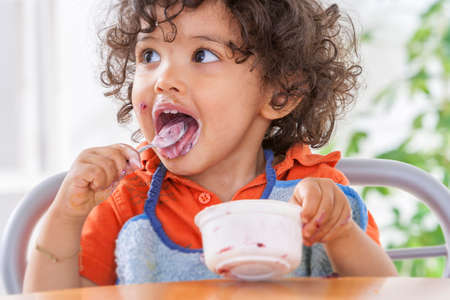 Toddler sitting in highchair and eating greek yogurt. Baby learning to eat and has yogurt on face and hairの写真素材