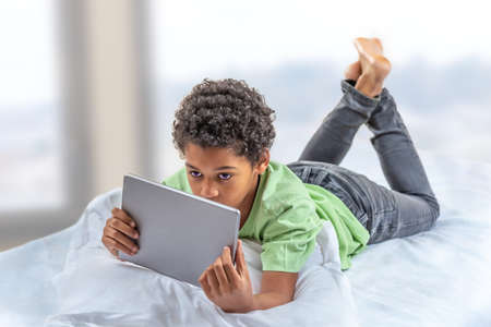 Boy teenager with tablet computer lying in bed having fun in bedromの写真素材
