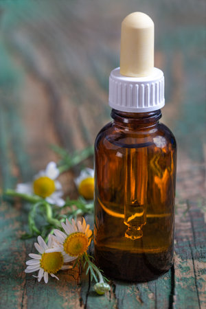 Small glass bottle with essential roman chamomile oil on the old wooden background. Chamomile flowers, close up. Aromatherapy, spa and herbal medicine ingredients. .の写真素材