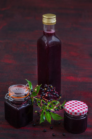 Homemade black elderberry syrup in glass jar and bunches of black elderberry with green leaves on wooden desk. Herbal medicine.の写真素材