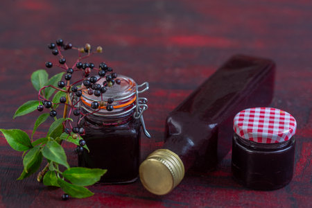 Homemade black elderberry syrup in glass jar and bunches of black elderberry with green leaves on wooden desk. Herbal medicine.の写真素材