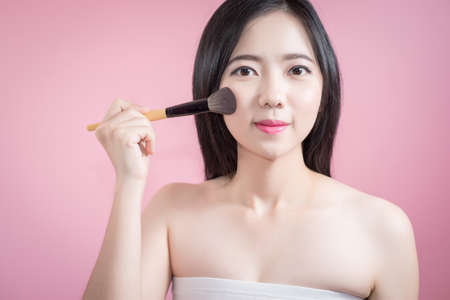 Long hair asian young beautiful woman applying cosmetic powder brush on smooth face isolated over pink background. natural makeup, SPA therapy, skincare, cosmetology and plastic surgery conceptの写真素材