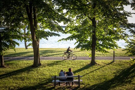 PALICE, SERBIA - JULY 1, 2018: Cyclist passing by on the coast of  Palic Lake with people sitting on a bench in Subotica, Serbia, with a green lawn in the foreground, during sunset

Picture of  the waters of the Palic Lake during a summer evening at dusk,のeditorial素材