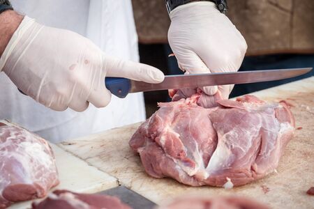 Hands of a professional butcher cutting a piece of raw pork meat with a long knife, on a white plank, at a rural butchery of Serbia. Pork meat is one of the biggest productions in Serbia.Picture of a big piece of raw pork meat being cut and chopped on a white wooden plank in the countryside of Serbia by a professional rural butcher.の写真素材