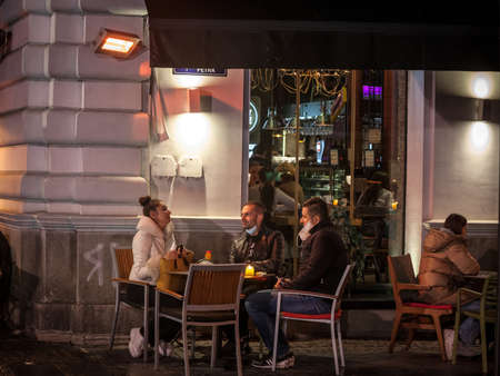 BELGRADE, SERBIA - OCTOBER 20, 2020: Three friends, clients at night at a heated terrace of a cafe serving food and drink wearing a respiratory face mask during the coronavirus covid 19 health crisis. Picture of a white caucasian people, clients of a cafeのeditorial素材