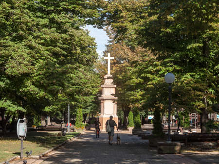 PANCEVO, SERBIA - SEPTEMBER 26, 2020: People passing by the calvary and catholic cross of the Trg Kralja Petra I square, the main square of Pancevo, one of the main cities of Banat region.のeditorial素材