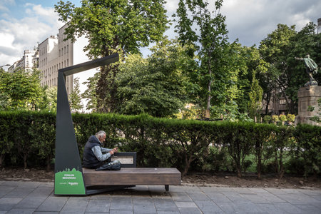 BELGRADE, SERBIA - MAY 24, 2020: Old senior man using a public phone charging station on a bench in a park to charge his smartphone in electricity while using internet.のeditorial素材