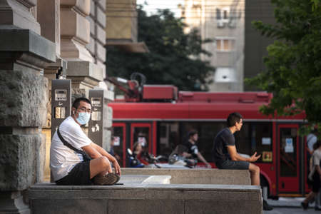 BELGRADE, SERBIA - AUGUST 4, 2021: Young Asian man, Chinese, tourist, wearing a respiratory face mask sitting in the streets of Belgrade.のeditorial素材