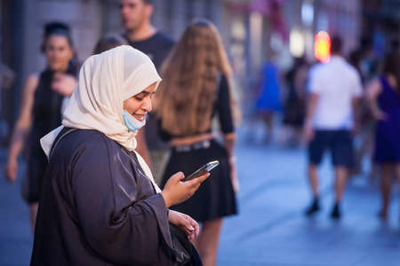 BELGRADE, SERBIA - JULY 22, 2021: Arabic Middle eastern young woman wearing islamic scarf veil wearing a face mask under the chin, during covid 19 coronavirus crisis, using her smartphone.,のeditorial素材