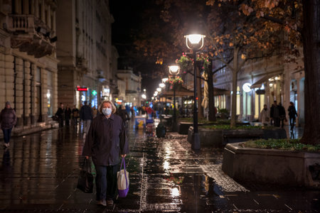 BELGRADE, SERBIA - NOVEMBER 3, 2021: Selective blur on an Old senior woman wearing a respiratory facemask walking at night with a groceries bag and shopping cart during coronavirus covid 19 crisis.のeditorial素材